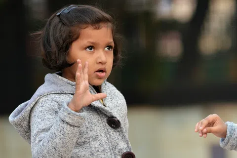 Two young girls in gray coats engaged in conversation with expressive gestures outdoors.