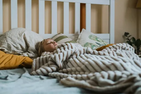 Young child sleeping peacefully under a soft, chunky knit blanket on a cozy bed with pillows.