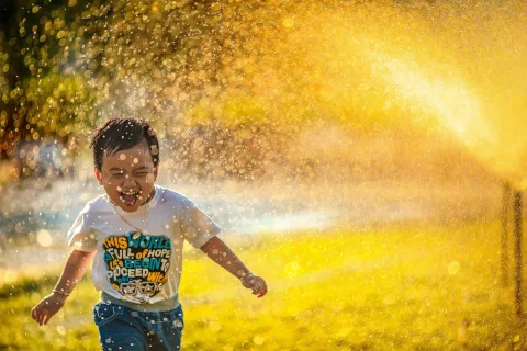 Joyful child runs through golden water spray on sunny day, laughing and playing outdoors with wet clothes.