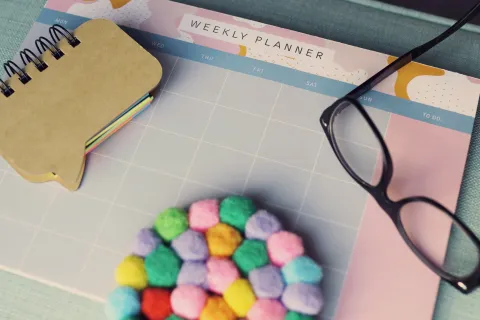 Weekly planner with blank grid, colorful pompom coaster, black glasses, and speech bubble sticky notes on blue surface