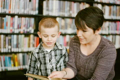 Woman reading a book to a young boy in a library with bookshelves in the background