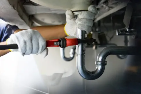 Plumber repairing pipes under a sink with a wrench and gloves.