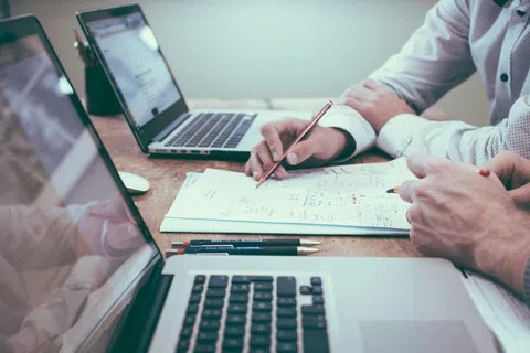Two people collaborate over documents and laptops discussing work details in a modern office setting.