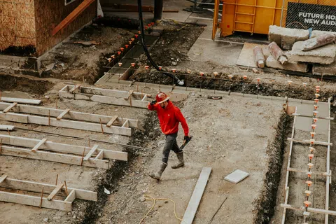 Construction worker in a red jacket and hard hat walking across a foundation site with gravel and wooden frames.