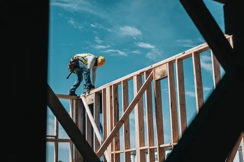 Construction worker wearing a safety helmet working on framing a wooden house under a clear blue sky.