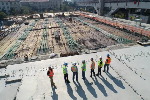 Construction workers in safety gear inspecting a large building site with exposed pipes and wiring beneath a concrete slab.