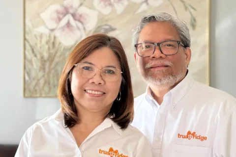 Smiling man and woman wearing white trustbridge shirts stand in front of floral wall art indoors.