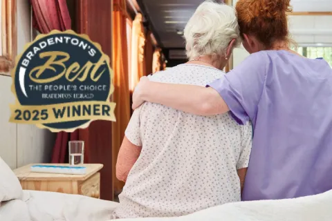 Caregiver in purple scrubs embraces elderly woman sitting on bed in cozy room with 2025 winner badge.