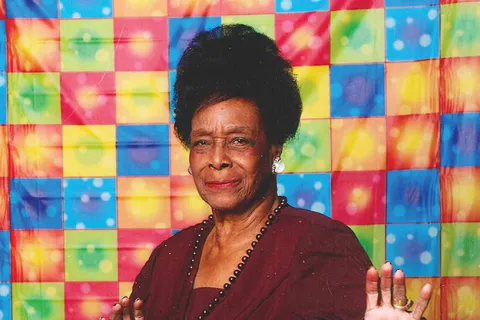 Dr. Bernice Catherine Harper in maroon outfit posed with hands raised against colorful checkered backdrop.