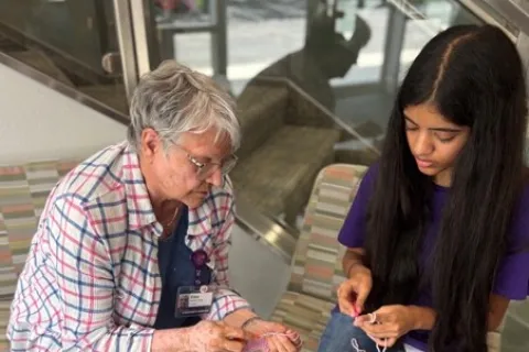 Older woman teaching young girl to crochet while seated on striped chairs with yarn containers on the floor.