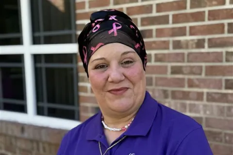 Smiling woman in a purple Hospice of Marion County polo and pink ribbon headscarf stands outside a brick building.
