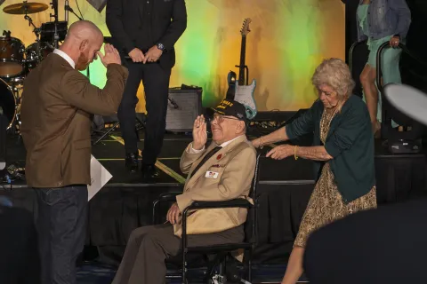 Veteran in wheelchair salutes man standing nearby as elderly woman pushes wheelchair at event stage with musical instruments.