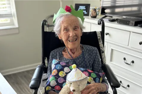 Elderly woman in a wheelchair wearing a green paper crown, smiling and holding a teddy bear with a party hat.