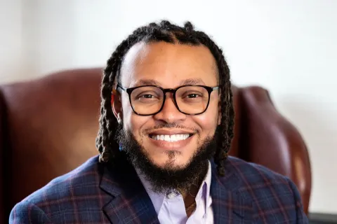 Smiling man with glasses and beard wearing a blue plaid blazer and white shirt seated in a leather chair