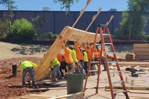 Construction workers in safety gear lifting and positioning a wooden frame on a sunny construction site.