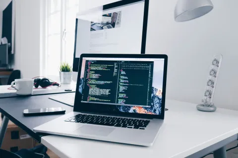 Laptop displaying code on a desk with a smartphone, headphones, coffee cup, and a large monitor in the background.