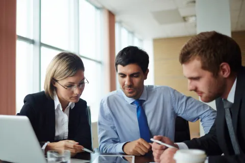 Three business professionals in formal attire collaborating around a tablet in a modern office.