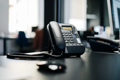 Black office desk phone with keypad and coiled cord on a dark modern office desk with blurred background