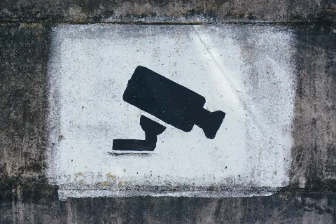 Black surveillance camera symbol painted on a weathered concrete wall with white background patch.