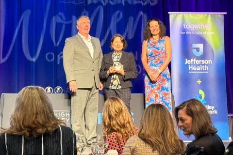 Three people standing on stage at a business women awards event with Jefferson Health banner and audience in foreground
