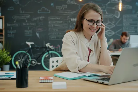 Woman wearing glasses talks on phone while working on laptop in creative office with bike and chalkboard.
