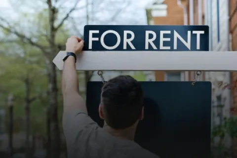 Person hanging a For Rent sign outside a residential property on a tree-lined street.