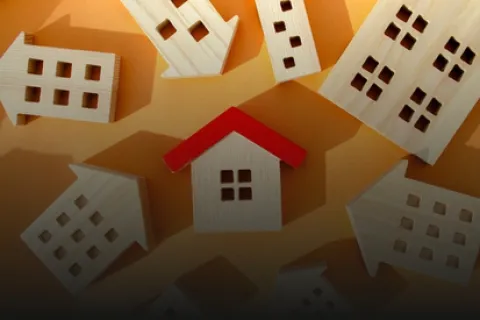 Wooden house models with one red roof highlighted among others on an orange background.