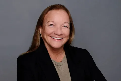 Smiling woman in a black blazer and beige shirt posing with arms crossed against a gray background
