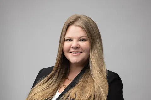 Smiling woman with long blonde hair wearing a black blazer against a gray background