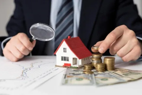 Person in suit examining small house model with magnifying glass and stacking coins on dollar bills