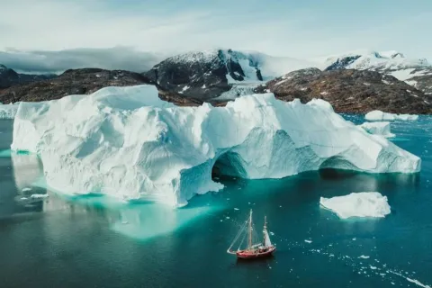Large iceberg with a tunnel and icy mountains in the background, a sailing ship in turquoise Arctic waters.