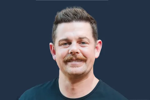Smiling man with styled brown hair and mustache wearing a black shirt against a dark blue background