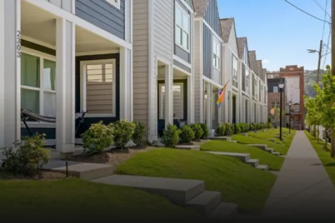 Row of modern townhouses with porches, manicured lawns, and a sidewalk under a clear blue sky.