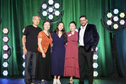 Five people standing together on stage with green backdrop and hexagonal LED lights, smiling at the camera