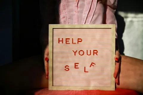 Person holding letter board with red text 'HELP YOURSELF' in sunlight with shadows and pink shirt.