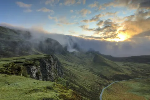 Sunset over misty green cliffs and winding road in a mountainous landscape under a cloudy sky