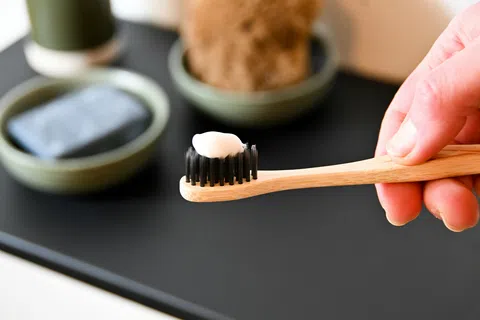 Hand holding a bamboo toothbrush with black bristles and white toothpaste on it over a black surface with soap and brown sponge.