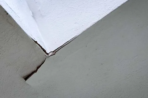 Close-up of a large crack at the corner of an interior wall with white and gray paint, showing structural damage.