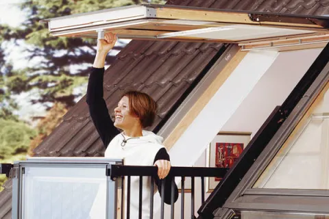 Woman standing on rooftop balcony with glass railing opening a large window on a sloped roof under clear sky