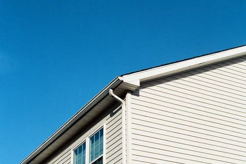 Upper corner of a beige house exterior with siding, windows, gutters, and clear blue sky background.