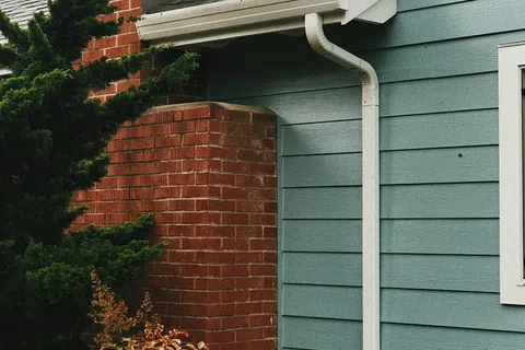 Close-up of a blue house exterior with white gutter, gray roof shingles, brick chimney, and green foliage.
