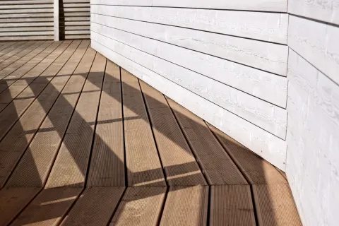 Sunlight casts geometric shadows on brown wooden deck with white horizontal siding wall.