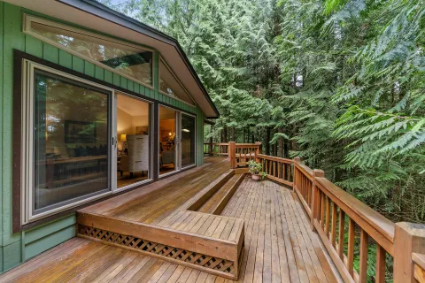Wooden deck with built-in seating attached to green house surrounded by tall trees and plants.