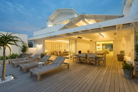 Modern outdoor patio with wooden lounge chairs, dining table, and warm indoor lighting at dusk.