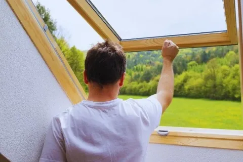 Man in white t-shirt opens a wooden framed skylight window with a scenic green landscape outside.