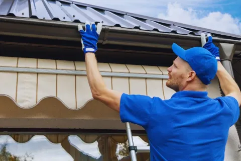 Professional worker in blue uniform installing or cleaning a metal roof gutter on a sunny day.
