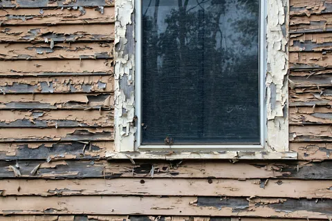 Old wooden house wall with peeling paint and a single dirty window reflecting trees and sky.