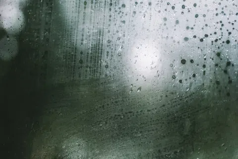 Close-up of rain droplets and streaks on a foggy glass window with blurred green and white background.