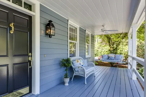 Cozy front porch with blue floor, white wicker chairs, floral cushions, black door, and surrounding greenery.
