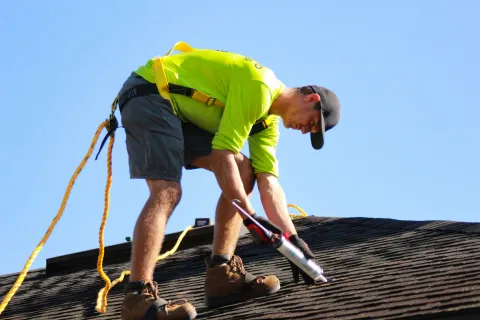 Construction worker in safety harness applying sealant on roof shingles under clear blue sky